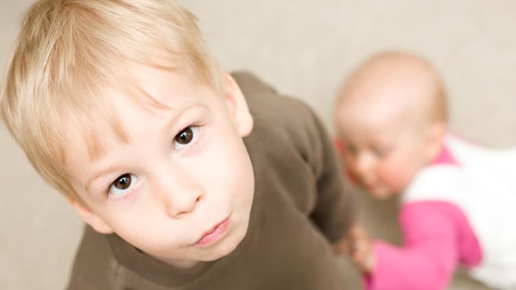Ein kleiner Junge schaut genervt, während ein Baby sich an ihm hochzieht.
