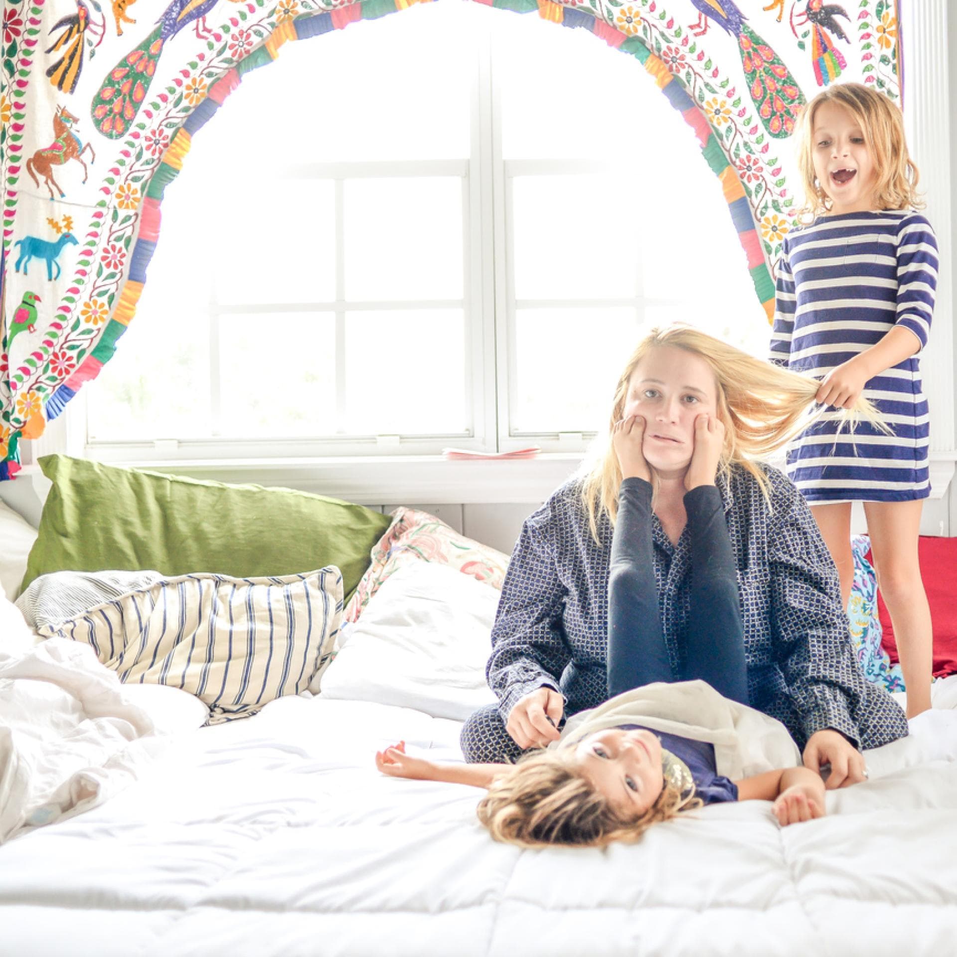 Ausgelaugte Mutter sitzt auf Bett, Kinder ziehen an ihren Haaren und machen Quatsch.
