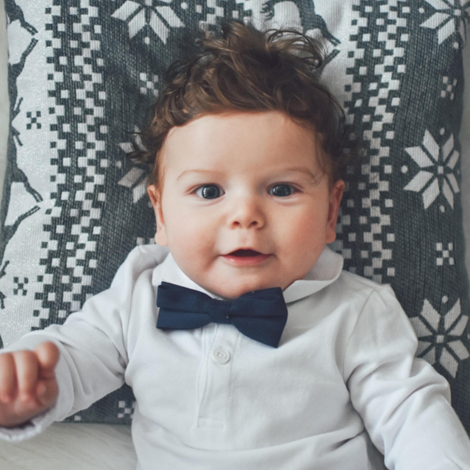 Kleiner Junge mit Fliege lächelt auf einem Kissen liegend in die Kamera.