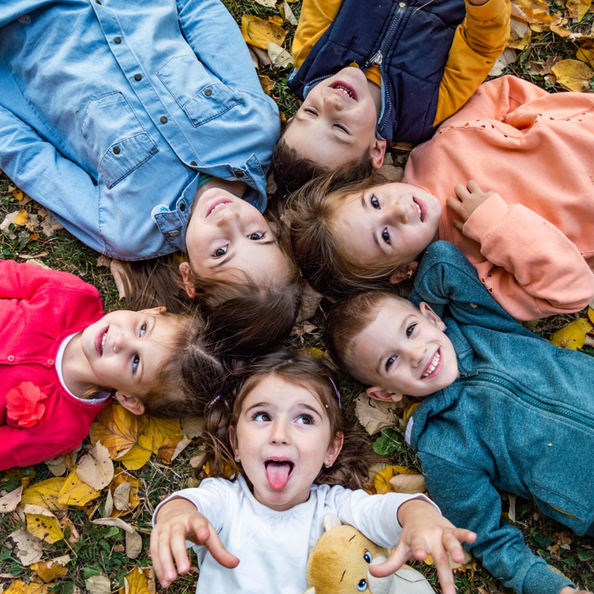 Kita-Kinder liegen im Herbstlaub und singen Herbstlieder im Kindergarten. 
