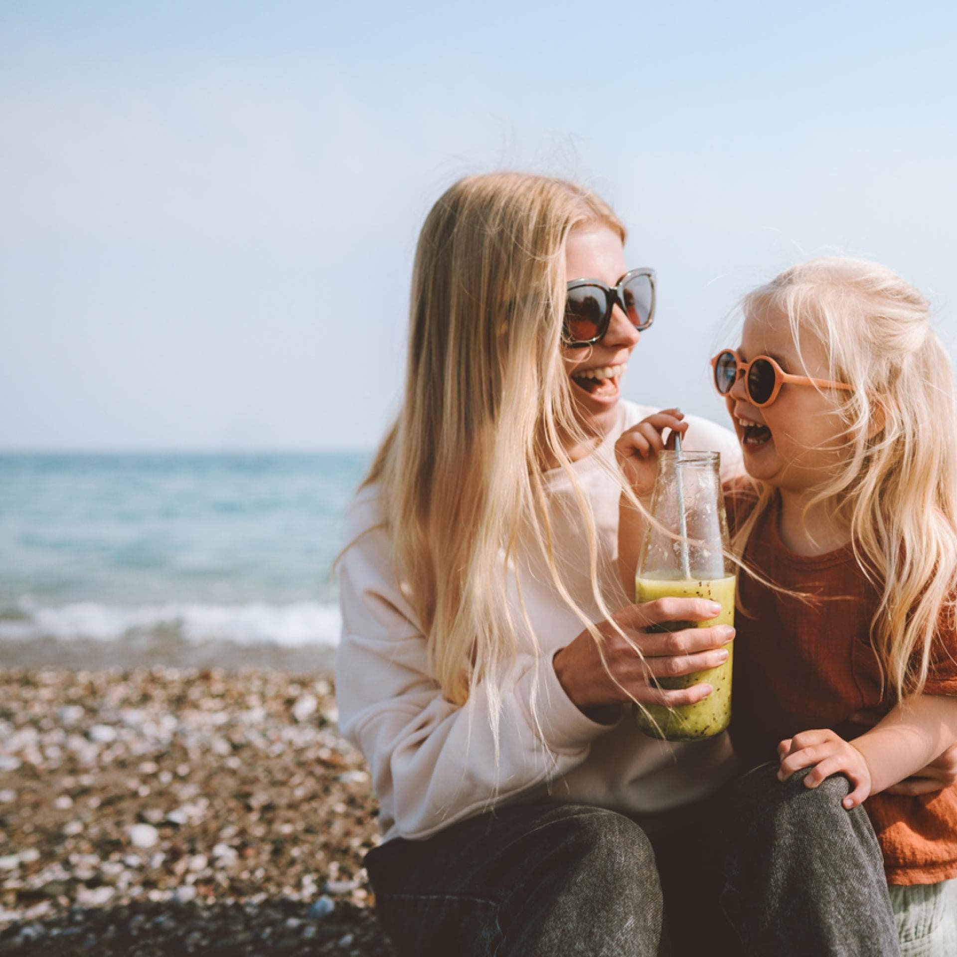 Mutter und Tochter sitzen fröhlich am schwedischen Strand, trinken einen Smoothie und lachen. 