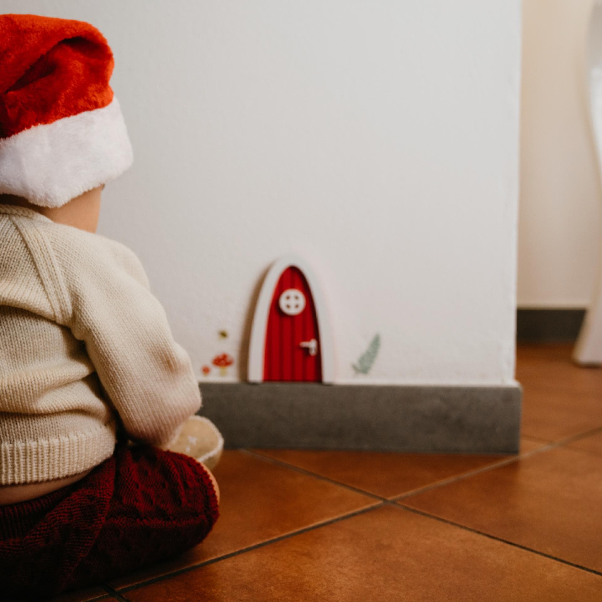 Baby-Junge mit Weihnachtsmütze sitzt vor der weihnachtlichen Wichteltür