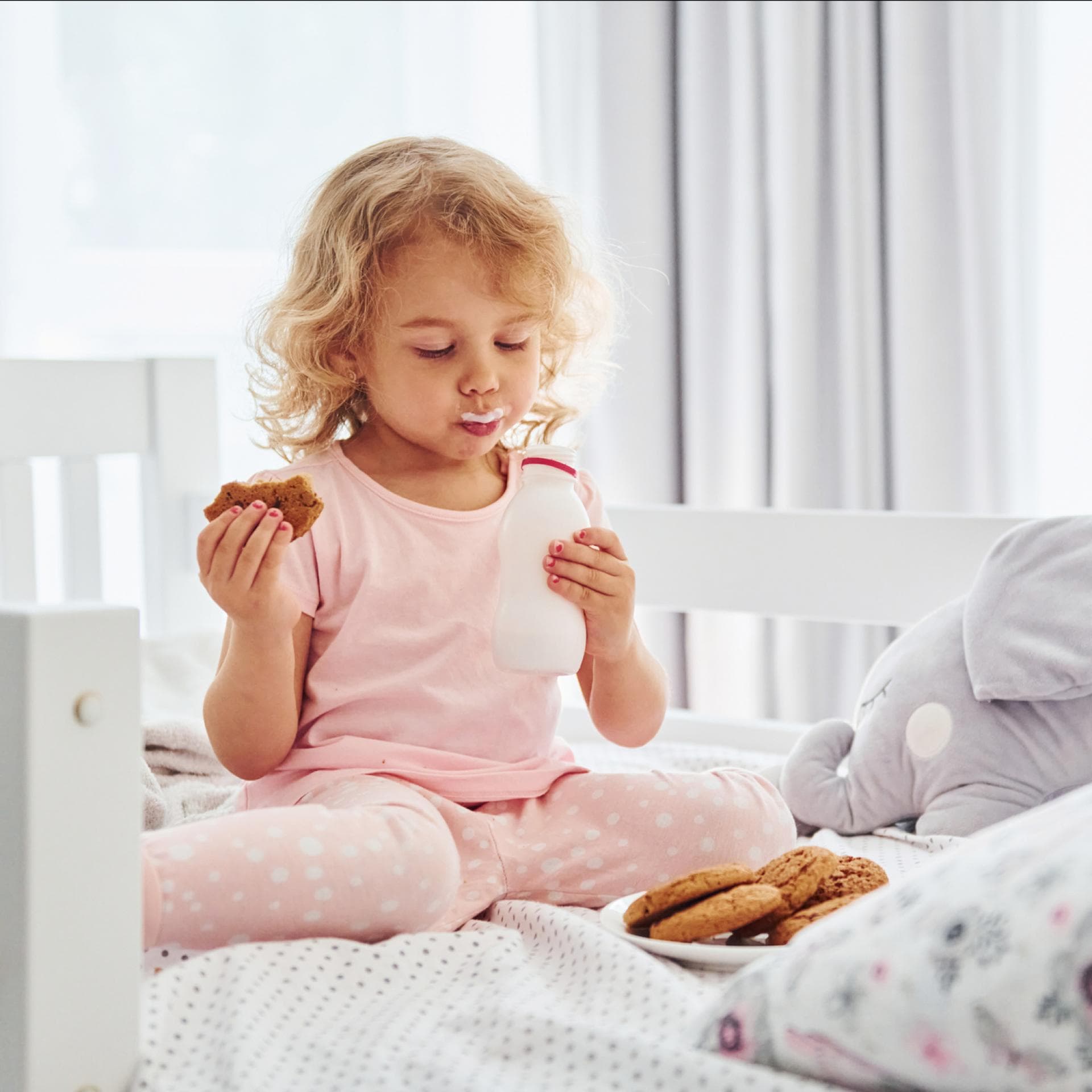 Ein kleines Mädchen isst im Bett Kekse und trinkt Milch.