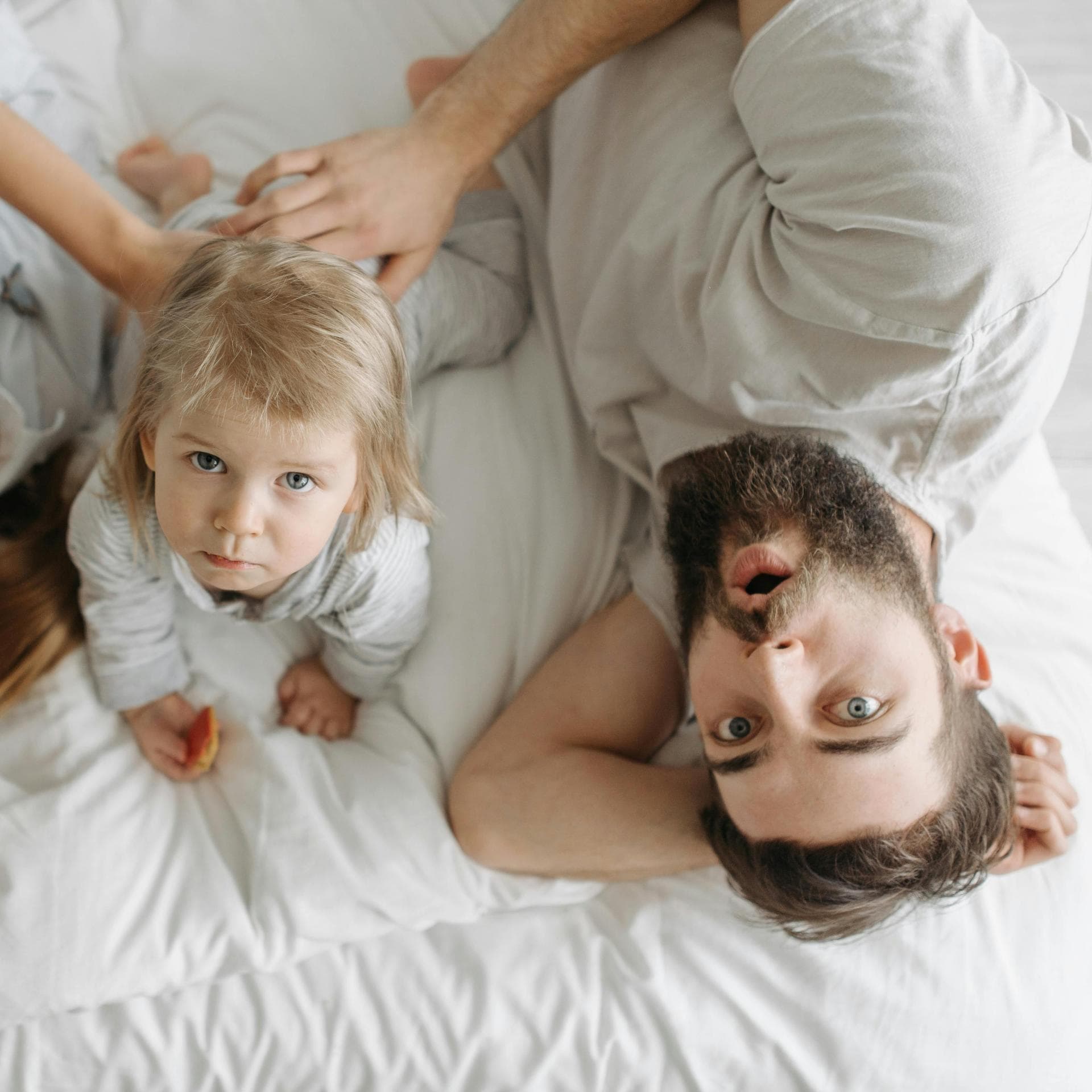 Eine glückliche Mutter liegt mit ihrem eher traurigen Kleinkind und neuen Partner im Bett.