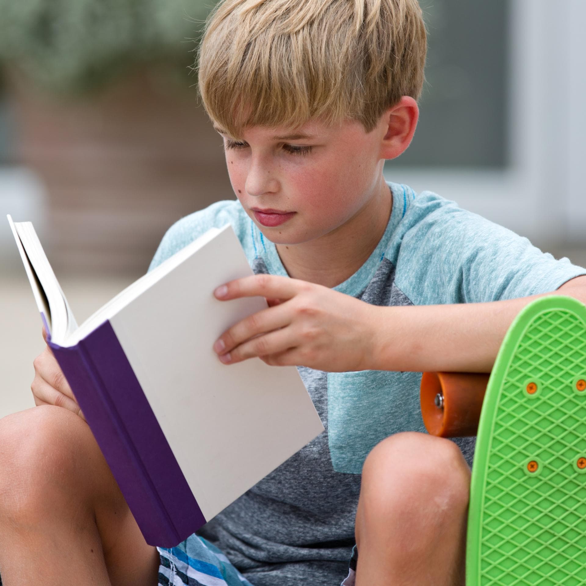 Junge mit Skateboard liest in einem Buch.