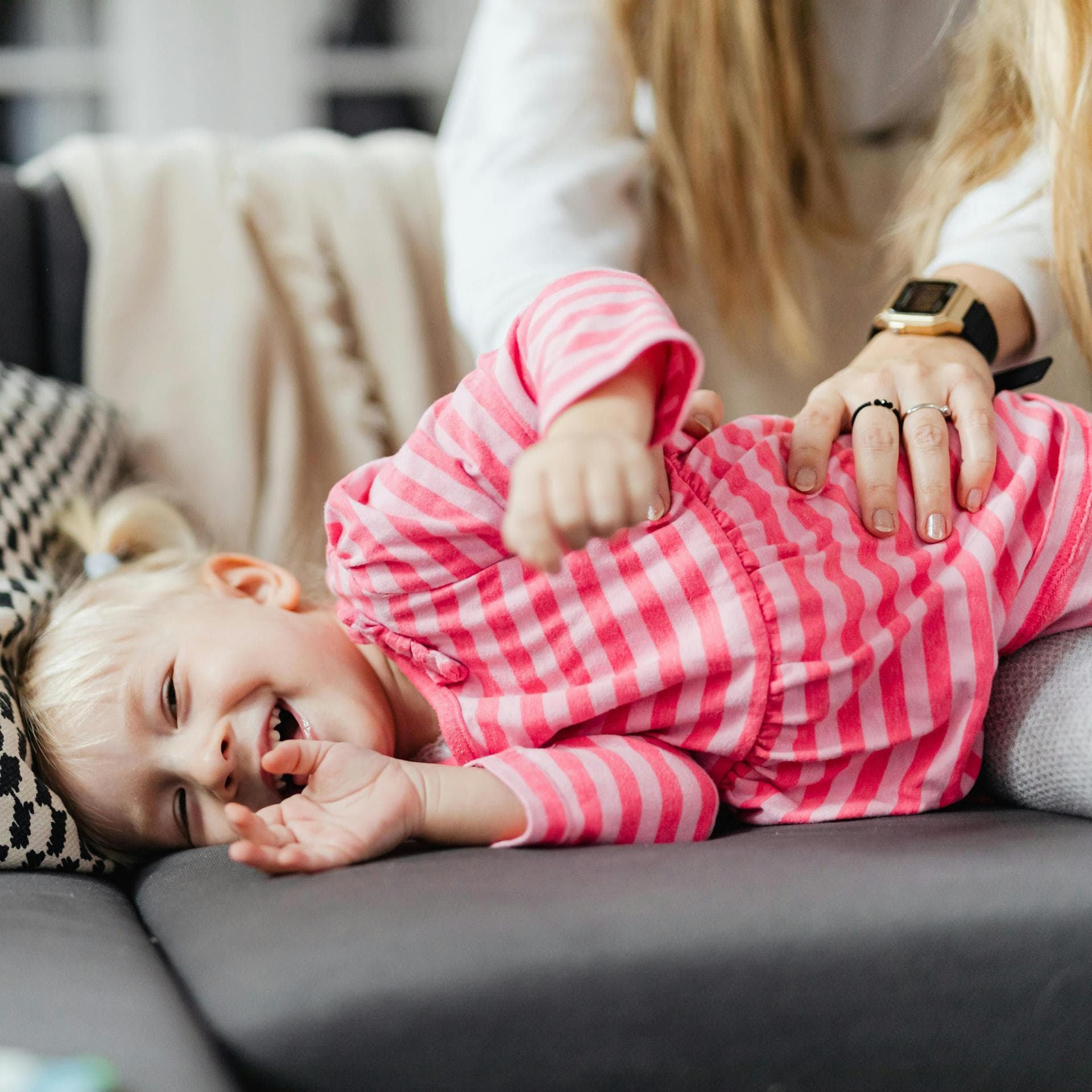 Ein kleines Mädchen wird von seiner Mutter auf dem Sofa gekitzelt.