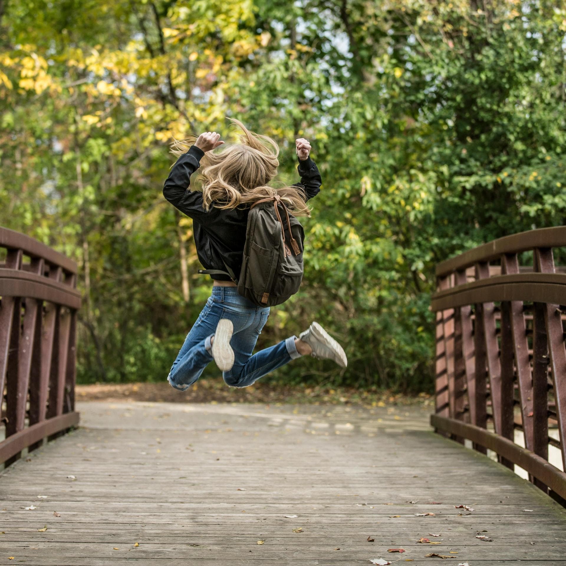 Mädchen mit Schulranzen springt hoch auf einer Holzbrücke.