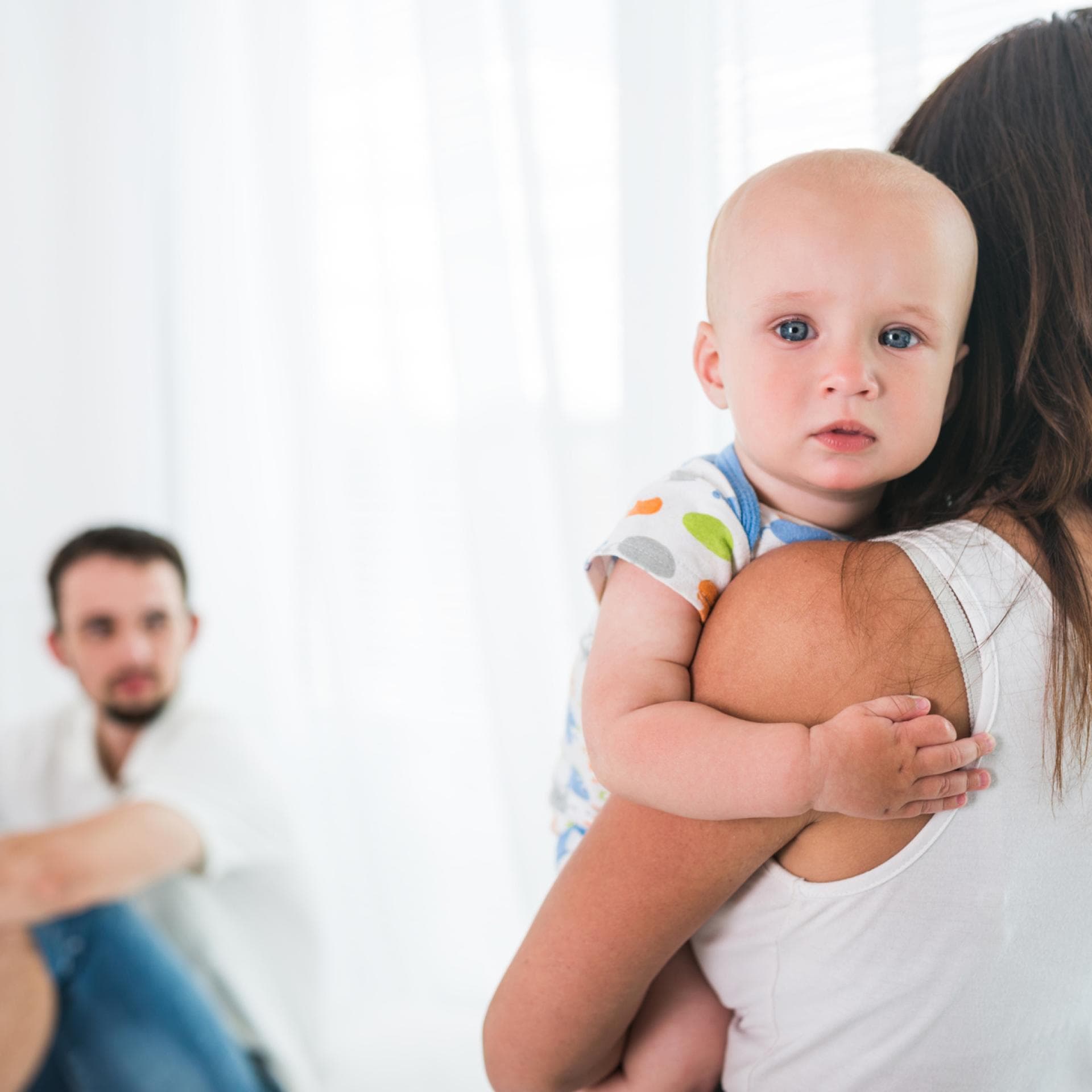 Mutter hält Baby auf dem Arm, Vater sitzt im Hintergrund.