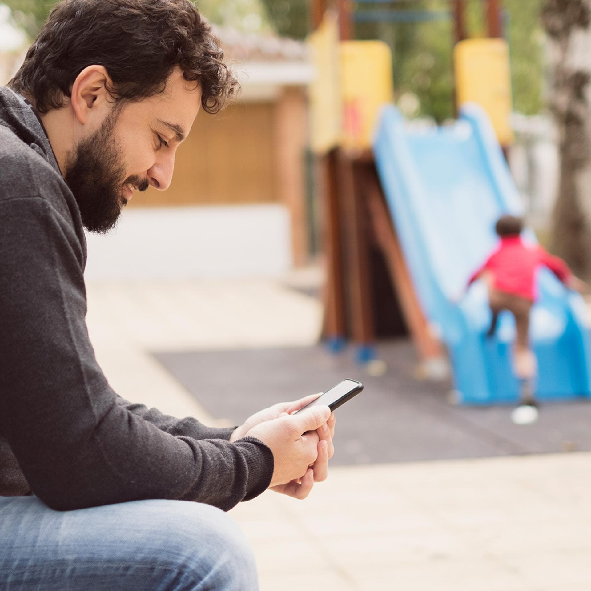 Vater mit Handy auf Spielplatz.