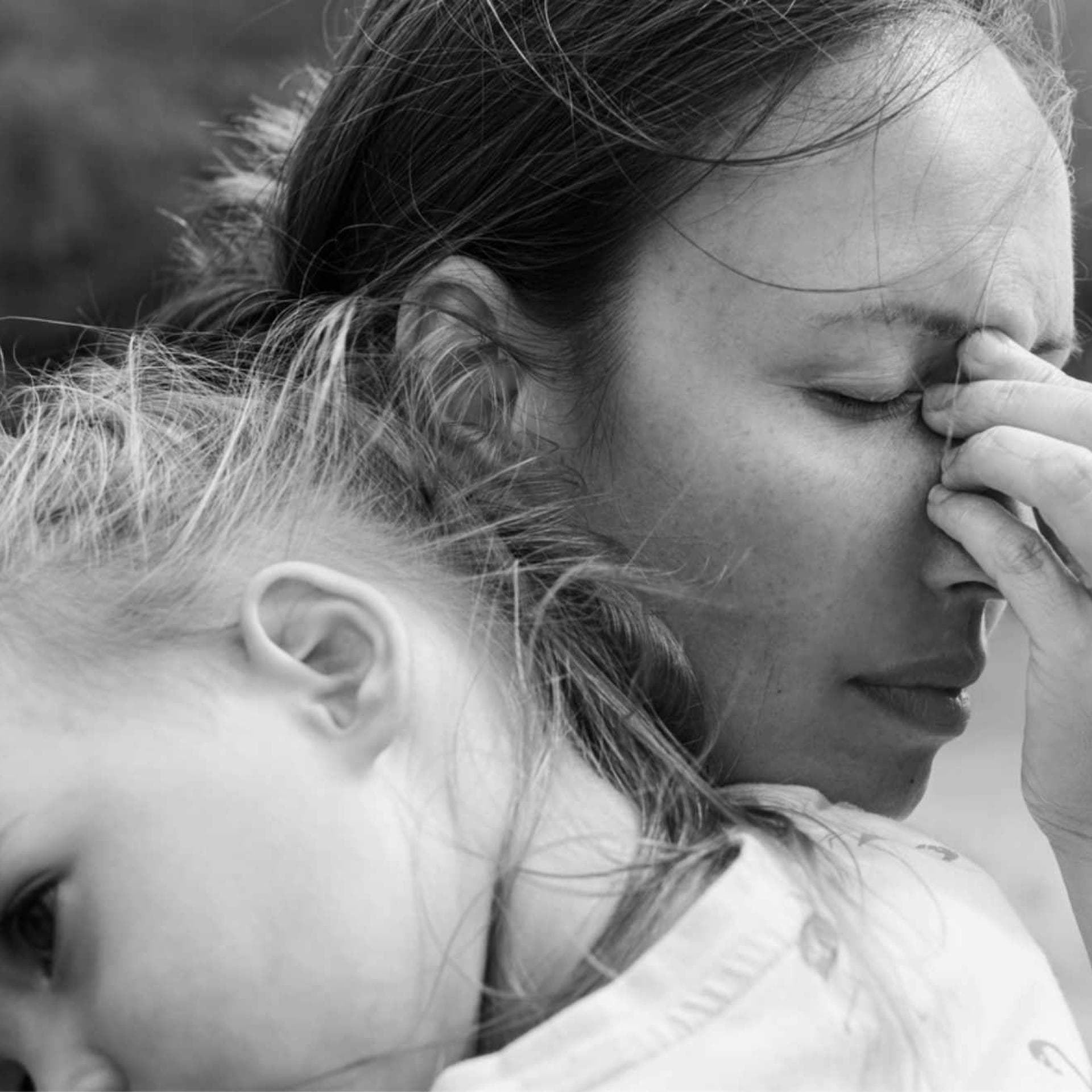 Eine verzweifelte Mutter hält ihre Tochter auf dem Arm, während sie die Stirn runzelt, die Augen schließt und sich an die Nase fässt.