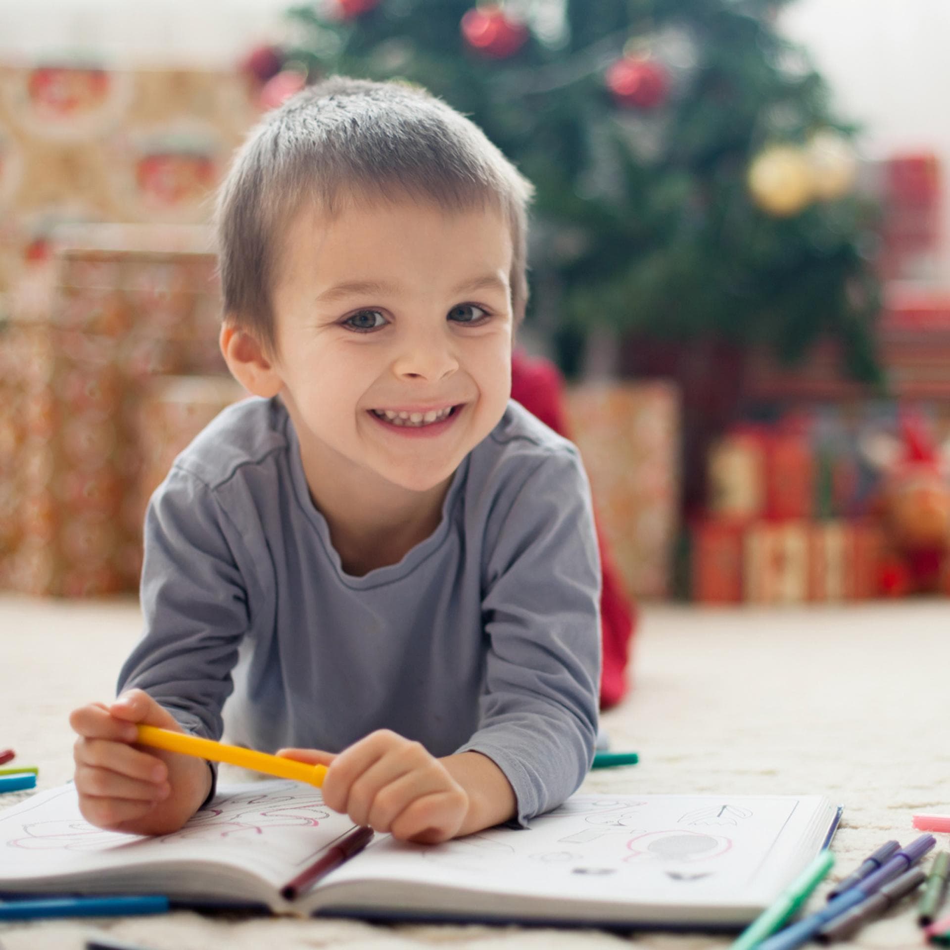 Ein kleiner Junge liegt vor dem Weihnachtsbaum auf dem Boden und malt Bilder..