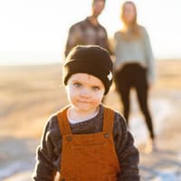 Ein kleiner Junge mit Wollmütze und Latzhose, der mit seinen Eltern am Strand ist.