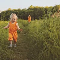 Ein kleines Mädchen in orangenem Sommeroutfit läuft durch eine Wiese.