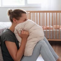 Eine Frau schreit in ein Kissen, während ihr Baby im Bett schläft
