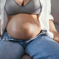 Eine Frau mit Babybauch sitzt mit geöffneter Hose auf dem Bett
