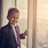 Ein Junge steht im Anzug am Fenster mit einer Kaffeetasse. 