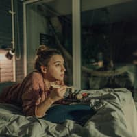 Eine Frau liegt abends im Bett, schaut TV und isst Sushi
