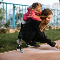 Babysitterin malt mit bunter Straßenmalkreide; ein Mädchen umklammert sie von hinten und schaut zu.