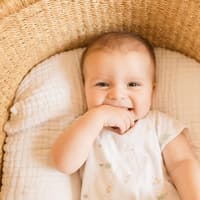 Ein kleiner, süßer Junge, der seine Finger im Mund hat und vom Babybett in die Kamera grinst.