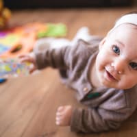 Kleines Baby-Mädchen mit Haarband liegt auf dem Boden und grinst.