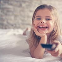 Lachendes Mädchen liegt mit Fernbedienung im Bett.