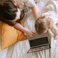 Frau und Kleinkind sitzen auf dem Bett und schauen auf einen Laptop, daneben ein Spiel-Tablett.