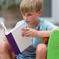 Junge mit Skateboard liest in einem Buch.