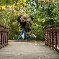 Mädchen mit Schulranzen springt hoch auf einer Holzbrücke.
