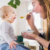 Mama füttert Baby mit Brei. Beide sperren den Mund auf.