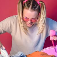 Fröhliche Frau mit pinker Brille bügelt und telefoniert gleichzeitig.