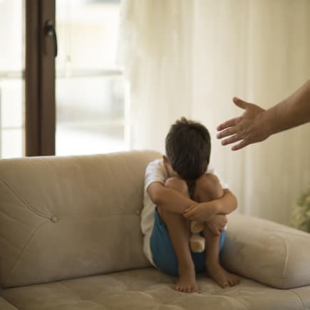 Junge sitzt mit angezogenen Knien auf dem Sofa, während sein Vater mit ihm schimpft.