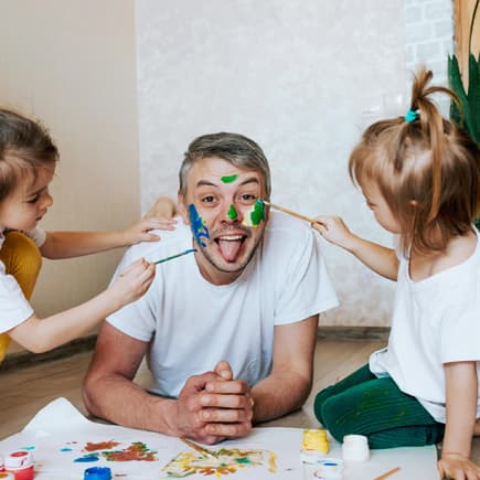 Ein männlicher Erzieher lässt sich in der Kita von zwei Kindern im Gesicht anmalen. 