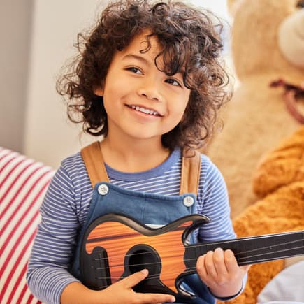 Ein Junge mit dunklen Locken sitzt mit seiner Gitarre im Kinderzimmer.