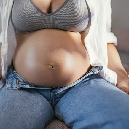 Eine Frau mit Babybauch sitzt mit geöffneter Hose auf dem Bett