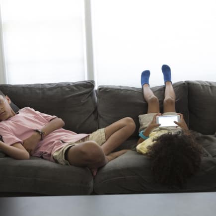Zwei Kinder liegen auf dem Sofa, eins mit Fernbedienung in der Hand, eins mit Smartphone