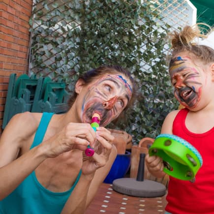 Eine junge Frau spielt mit einem Kleinkind, trällert auf der Flöte mit bemaltem Gesicht.