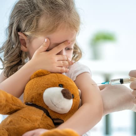 Kleines Mädchen mit Teddy im Arm bekommt eine Spritze in den Oberarm. 
