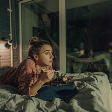 Eine Frau liegt abends im Bett, schaut TV und isst Sushi