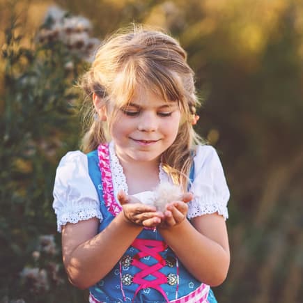 Ein kleines Mädchen im Dirndl steht auf dem Feld und hält eine Blüte in der Hand.