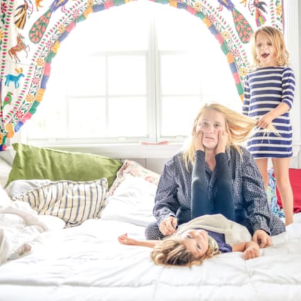 Ausgelaugte Mutter sitzt auf Bett, Kinder ziehen an ihren Haaren und machen Quatsch.