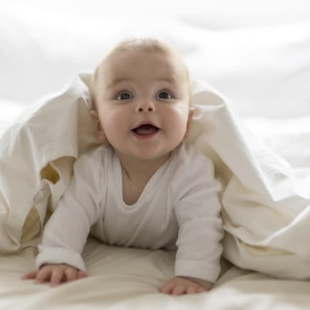 Ein kleiner Baby-Junge guckt frech unter einer weißen Bettdecke hervor. 