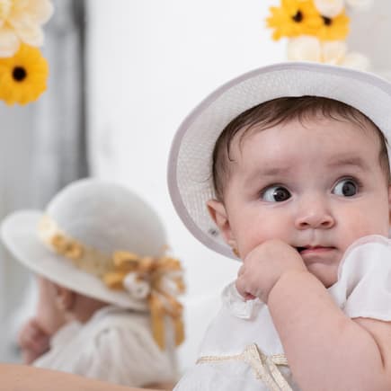 Ein kleines Baby-Mädchen mit Taufkleid und Hut schaut niedlich zur Seite. 