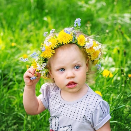 Kleines Mädchen mit Blumenkranz auf dem Haar steht auf einer Wiese.