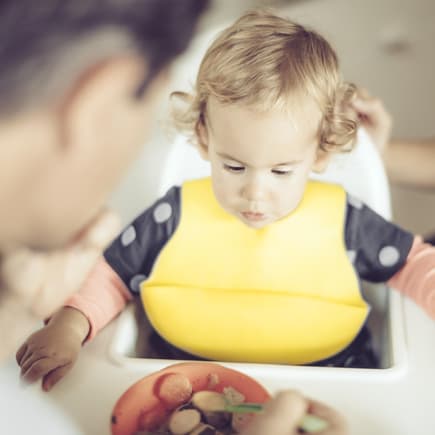 Baby-Mädchen bekommt ihr Essen im Hochstuhl – das Lätzchen verhindert großes Chaos