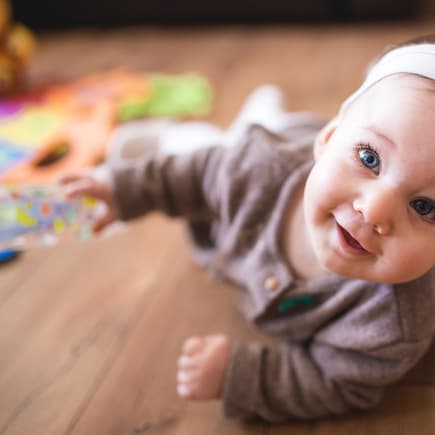 Kleines Baby-Mädchen mit Haarband liegt auf dem Boden und grinst.