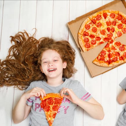 Zwei fröhliche Kinder liegen mit Salami-Pizza auf dem Boden.