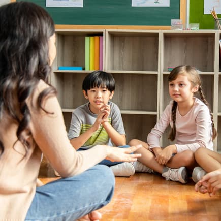 Lehrerin sitzt mit Grundschulkindern auf dem Boden.
