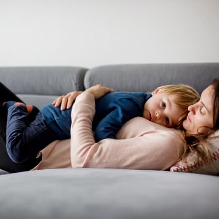 Mutter hält krankes Kind, liegen zusammen auf einer Couch. 