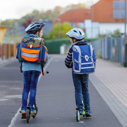Zwei Kinder mit Schulranzen auf dem Rücken fahren auf Rollern zur Schule.