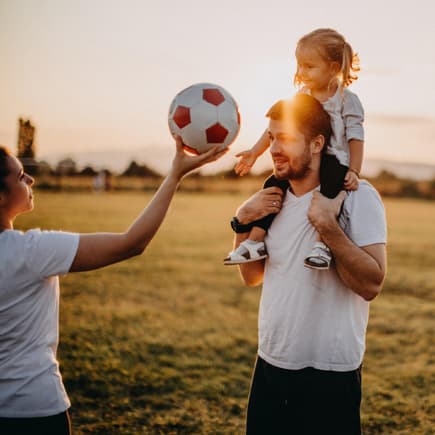 Mutter hält Mädchen einen Fußball hin, die auf Papas Schultern sitzt.